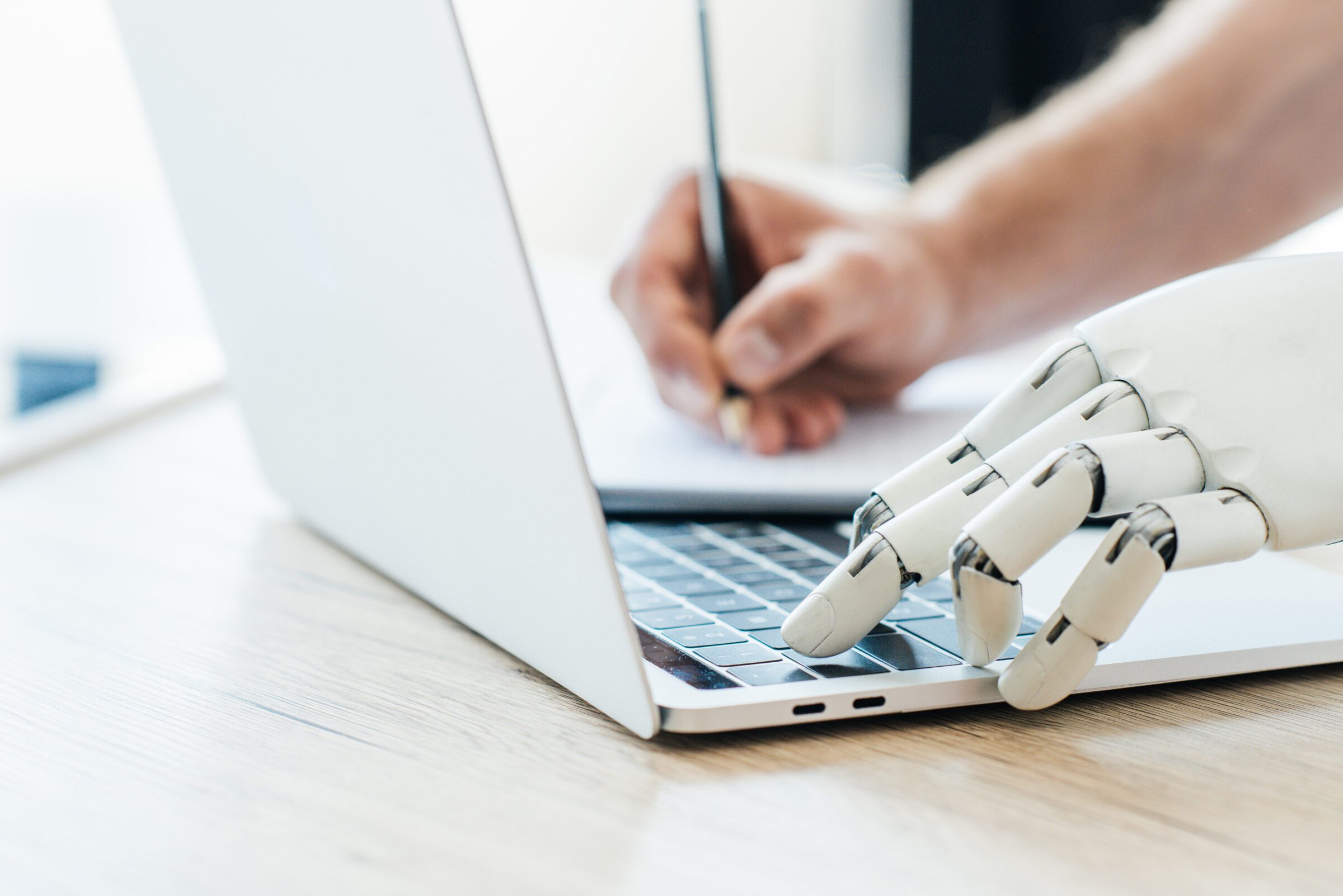 close up view of robotic arm using laptop and human hand taking notes at wooden table